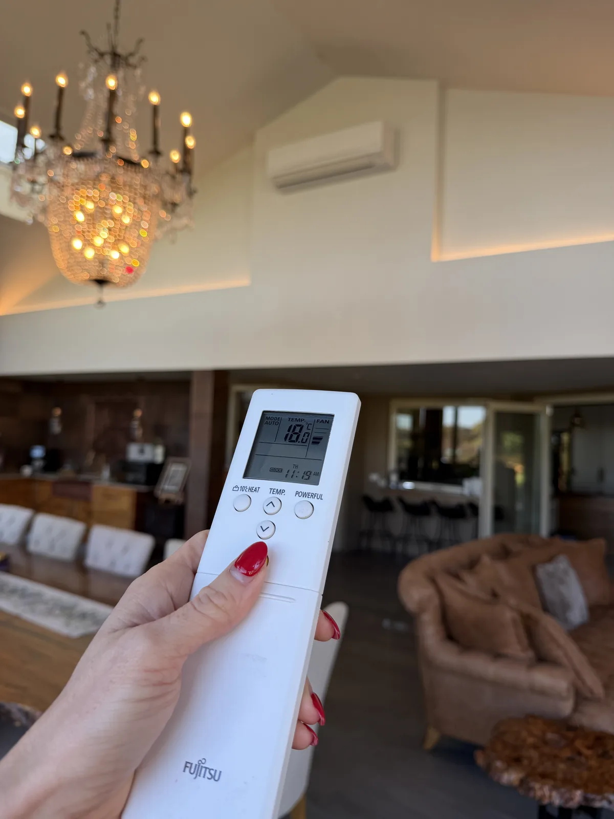 A hand holding an air-conditioner remote control beneath a wall-mounted split-system unit in a Perth home